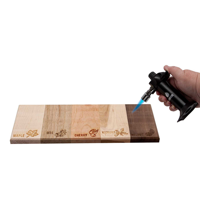 Hand holding a butane torch over a wooden smoking board with engraved wood type names.