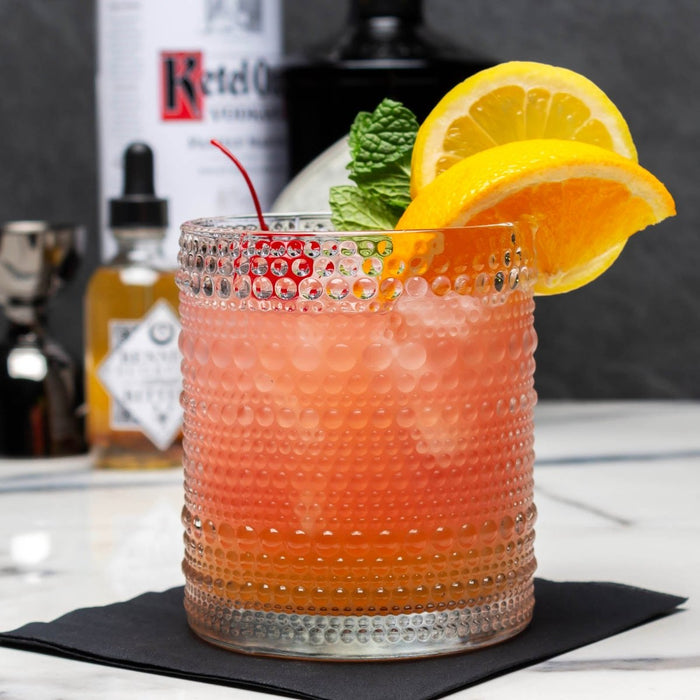Cocktail in a textured Jupiter Glass glass with orange slices and a cherry, with Ketel One vodka in the background.