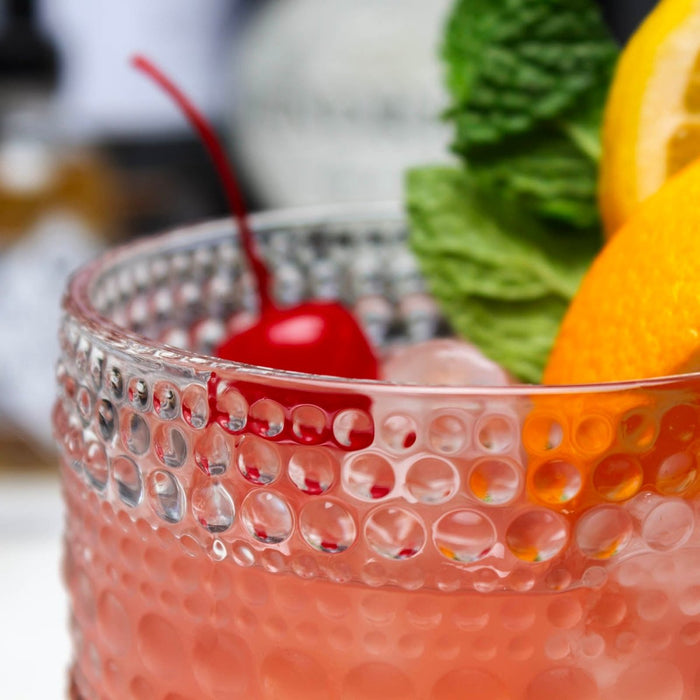 Close-up of a pink cocktail with a cherry, lemon, and mint leaf in a textured glass.