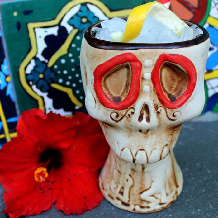 Skull-shaped mug with red glasses filled with a drink and ice, next to a red flower on a colorful background.
