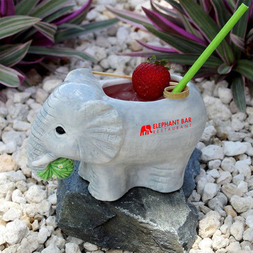 Elephant-shaped drink holder with a drink and strawberry on a pebbled surface with plants in the background