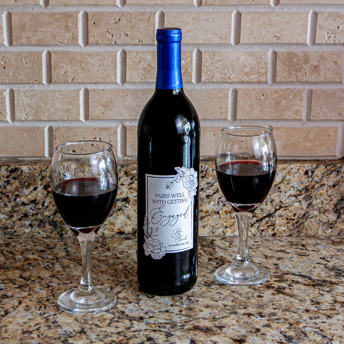 Wine bottle labeled 'Pairs well with getting Engaged' with two glasses of red wine on a countertop.