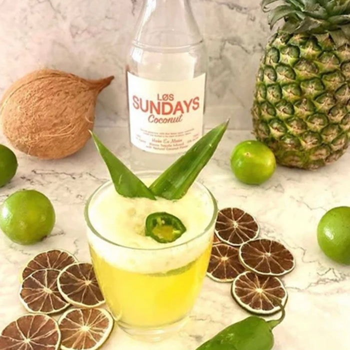Glass of yellow liquid with green leaves, bottle labeled 'Los Sundays Coconut', pineapple, coconuts, and limes on a marble surface.