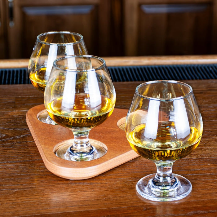 Three sniffer glasses on a cherry stain wooden tray with a blurred bar background
