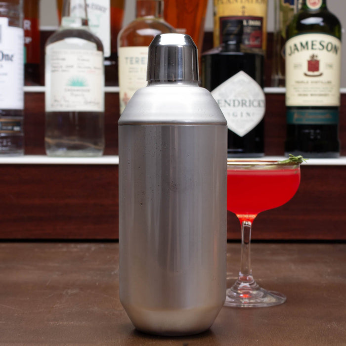 Metal cocktail shaker with a glass of red cocktail and alcohol bottles in the background