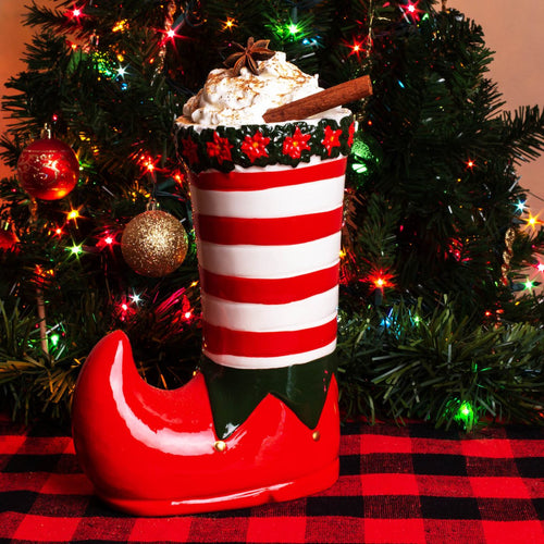 Decorative elf boot with a hot chocolate mug filled with whipped cream and a cinnamon stick, set against a Christmas tree background.