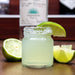 Mini mason jar with a shot of margarita with lime wedge on a wooden surface, with a bottle in the background.