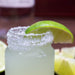 Mini mason jar with a shot of margarita with lime wedge on a wooden surface, with a bottle in the background.