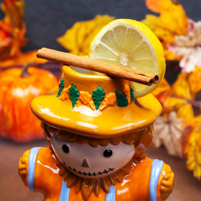 scarecrow tiki mug with a lemon slice and cinnamon stick on its head, surrounded by pumpkins.