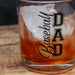 A clear rocks glass with a thick base, filled with an amber drink and ice, printed with the words 'Baseball DAD' in black. The glass is photographed close-up on a wooden surface.