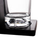 Clear glass tumbler with a thick base resting on a black square coaster or stand, photographed against a white background.