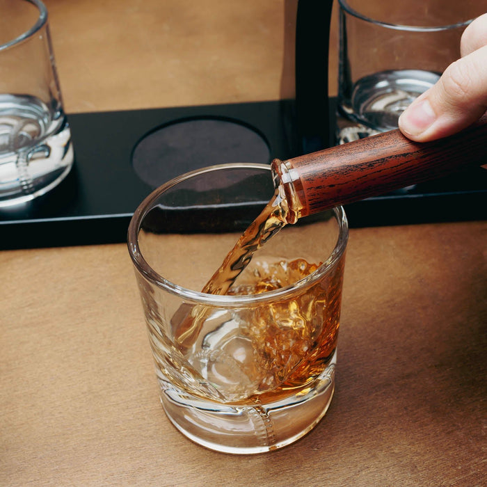 A clear cut-glass rocks tumbler with a textured pattern, being filled with amber whiskey from a decanter vessel held by a hand, set on a warm brown tabletop.