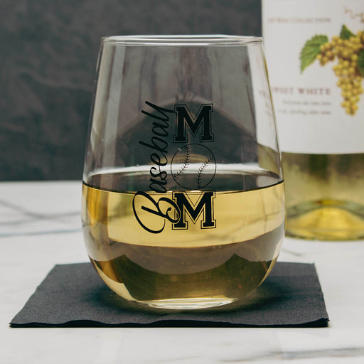 A clear stemless wine glass with black 'Baseball Mom', filled with white wine and resting on a dark napkin, with a wine bottle blurred in the background.