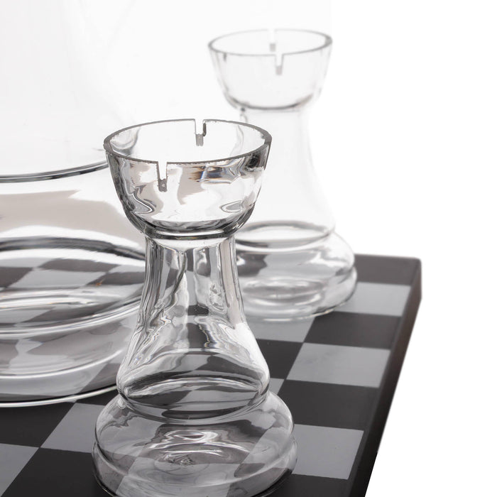 Clear glass chess-piece-shaped candle holders arranged on a black-and-white checkered surface against a white background.