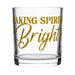 A clear glass rocks tumbler with gold foil lettering that reads 'Making Spirits Bright' on a solid white background.