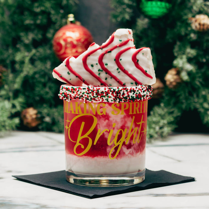 A festive holiday drink in a clear glass tumbler with gold script reading 'Taking Spirit to Bright,' topped with white whipped cream, red drizzle, and cookie slices, set on a black napkin with a blurred Christmas tree background.