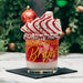 A festive holiday drink in a clear glass tumbler with gold script reading 'Taking Spirit to Bright,' topped with white whipped cream, red drizzle, and cookie slices, set on a black napkin with a blurred Christmas tree background.
