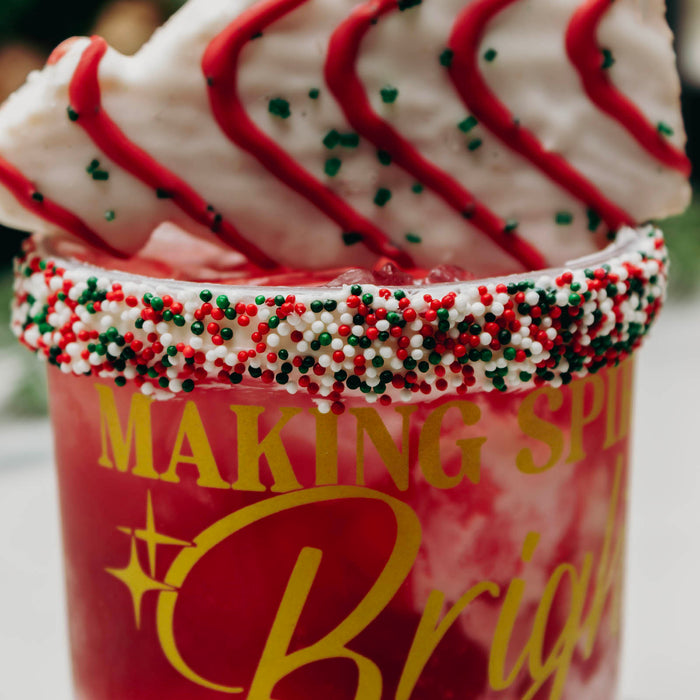 A festive red drink in a clear cup with yellow lettering, decorated with red, white, and green sprinkles around the rim, topped with a white pastry drizzled in red icing and sprinkles.