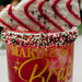A festive red drink in a clear cup with yellow lettering, decorated with red, white, and green sprinkles around the rim, topped with a white pastry drizzled in red icing and sprinkles.