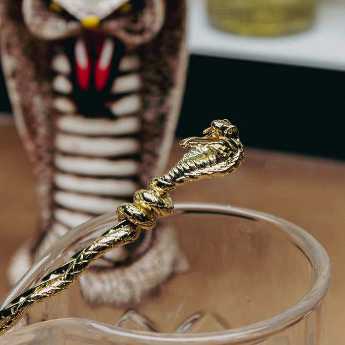 A close-up of a gold-toned snake-shaped spoon resting on a clear glass, with a blurred cobra tiki mug in the background.