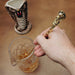 A hand holds a gold-toned metal spoon with a sculpted snake handle above a clear glass beaker containing amber liquid, with a small cobra tiki mug in the background on a tan surface.