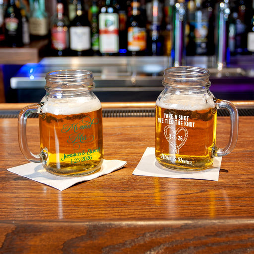 Two personalized clear glass mason jar mugs with handles, filled with beer, sitting on white napkins on a wooden bar counter with a blurred bar background.