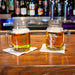 Two personalized clear glass mason jar mugs with handles, filled with beer, sitting on white napkins on a wooden bar counter with a blurred bar background.