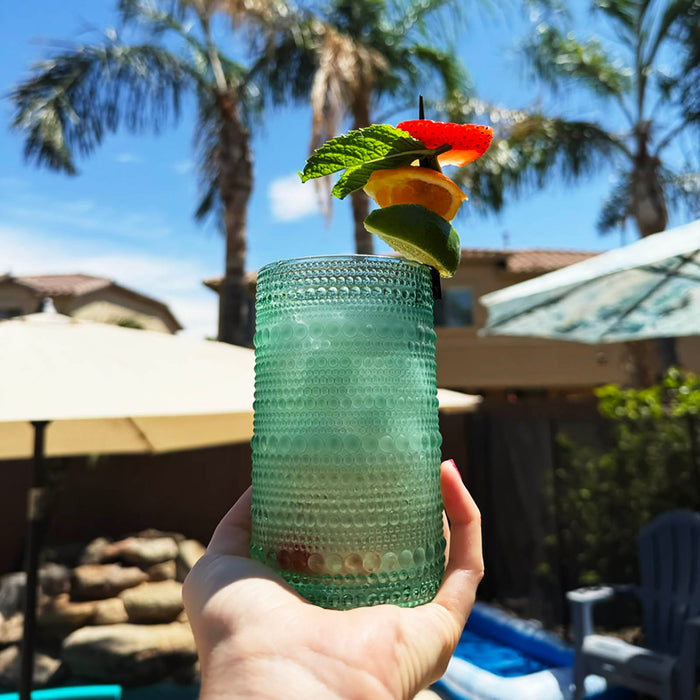 glass with a cocktail garnish held in front of a poolside setting with palm trees.