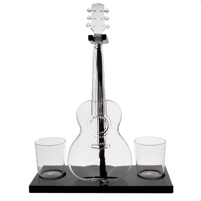 A clear glass acoustic guitar-shaped decanter with a black base, shown on a white background with two small clear glasses on either side of the base.