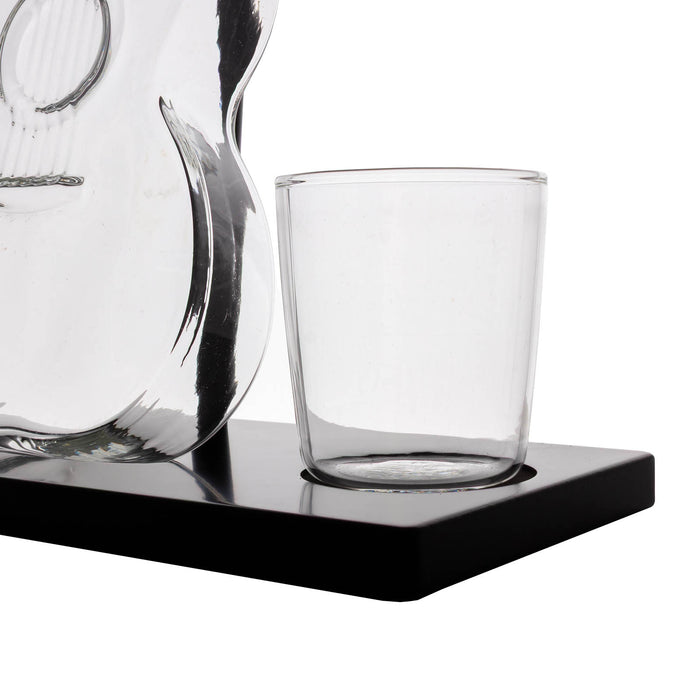 A clear glass tumbler cup sitting on a black rectangular base, with part of a transparent glass carafe visible at the left edge against a white background.