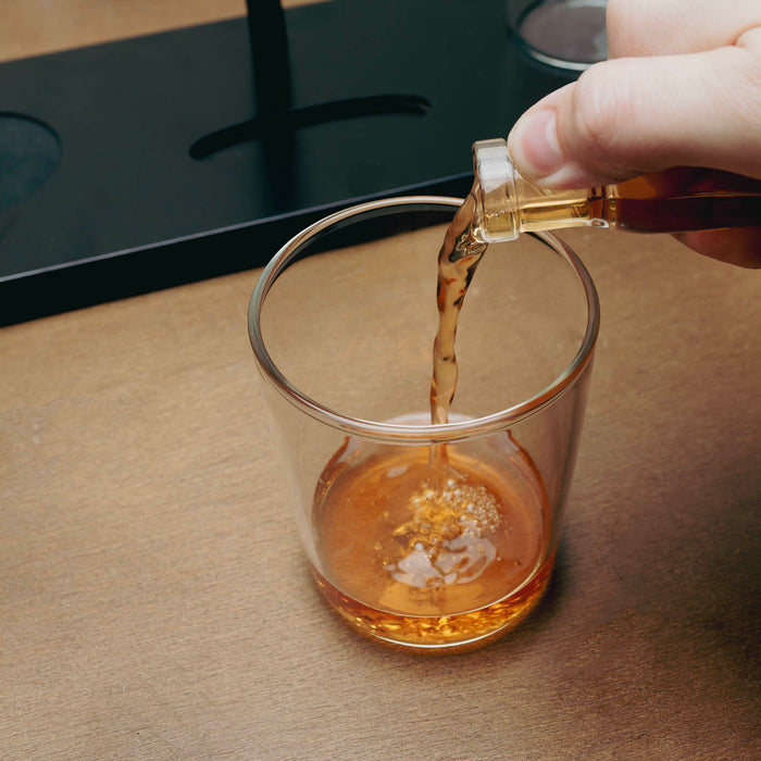 A hand pouring amber-colored liquid from a small clear bottle into a clear lowball glass on a wooden surface, with a dark tray in the background.