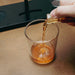 A hand pouring amber-colored liquid from a small clear bottle into a clear lowball glass on a wooden surface, with a dark tray in the background.