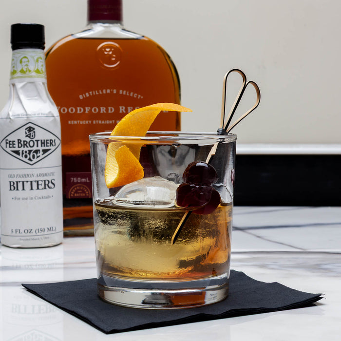 Glass of whiskey cocktail with garnishes on a table with bottles of Woodford Reserve and Fee Brother's Bitters in the background.