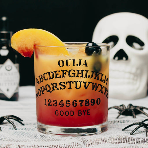 A clear glass tumbler printed with a Ouija board design, filled with a layered orange-to-red cocktail and garnished with a peach slice and black olives. The glass sits on a lace surface with a white skull, black spiders, and a dark bottle in the background.