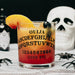 A clear glass tumbler printed with a Ouija board design, filled with a layered orange-to-red cocktail and garnished with a peach slice and black olives. The glass sits on a lace surface with a white skull, black spiders, and a dark bottle in the background.