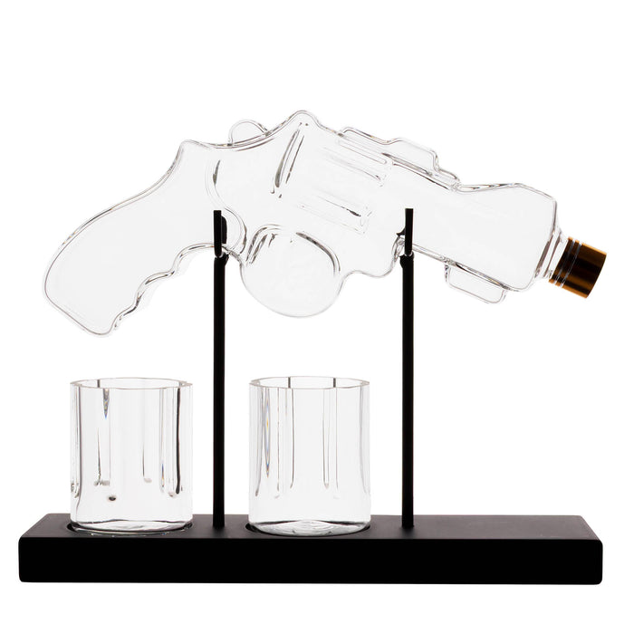 A clear glass revolver-shaped bottle holder on a black base with two small clear glasses, photographed against a plain white background.
