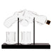 A clear glass revolver-shaped bottle holder on a black base with two small clear glasses, photographed against a plain white background.