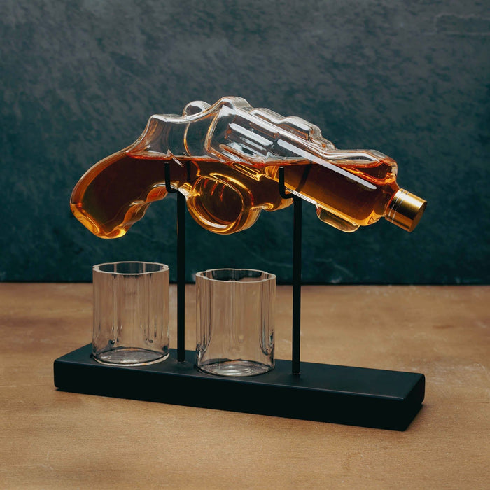 A clear glass revolver-shaped decanter filled with amber liquid, mounted on a black stand with two clear glasses below, set on a wooden surface against a dark textured background.