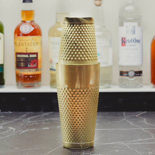 A gold-colored metal cocktail shaker with a textured, dimpled surface and a matching lid, standing on a dark marble countertop with blurred liquor bottles in the background.
