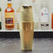A gold-colored metal cocktail shaker with a textured, dimpled surface and a matching lid, standing on a dark marble countertop with blurred liquor bottles in the background.