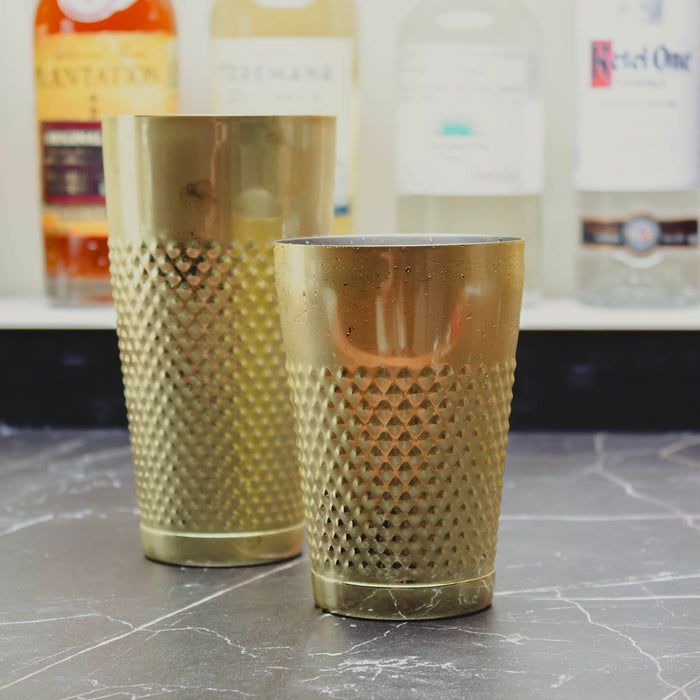 Two textured gold cocktail shakers with a diamond-patterned finish, one standing upright in the background and one sitting in the foreground on a dark marble countertop with blurred liquor bottles behind.