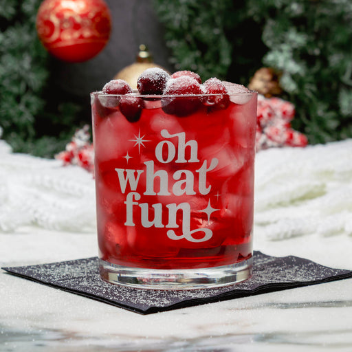 A clear rocks glass with white frosted lettering that reads 'oh what fun' sits on a black napkin. The glass is filled with a bright red drink and topped with cranberries, set against a blurred holiday background with greenery, ornaments, and white fabric.